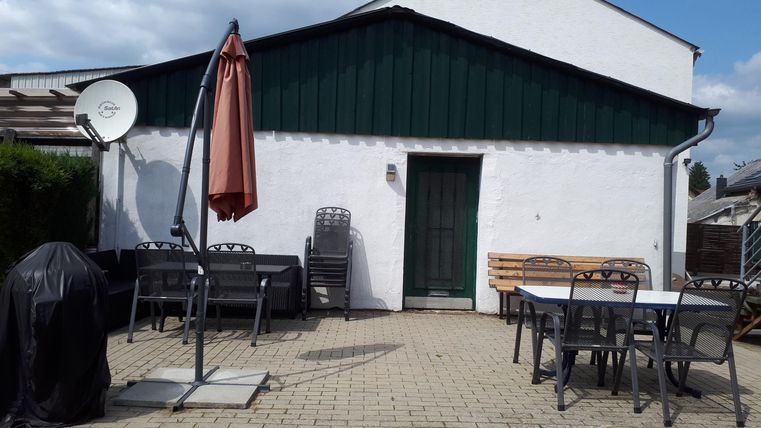 A cozy outdoor area with a table and chairs. In the background, there is a house with a green roof and an awning.