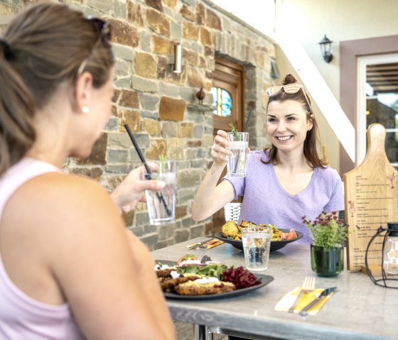 Zwei Frauen sitzen an einem Tisch im Freien, sto&szlig;en mit Getr&auml;nken an und genie&szlig;en eine Mahlzeit. Im Hintergrund eine Steinmauer., &copy; Eifel Tourismus GmbH, Dominik Ketz