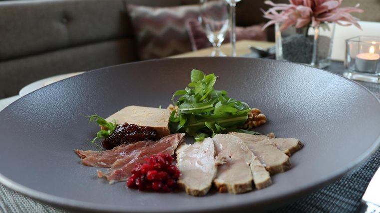Un plat élégant dans une assiette plate, composé de différentes viandes, de salade fraîche et d'un accompagnement de baies rouges. En arrière-plan, on peut voir une décoration de table raffinée et de la vaisselle en verre.