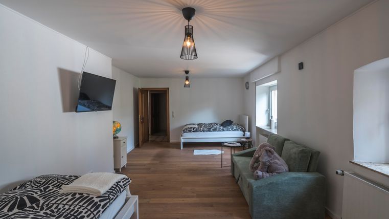 Ein helles Zimmer mit zwei Betten und einem gemütlichen Sofa. An der Wand hängt ein Fernseher, und der Boden ist aus Holz.