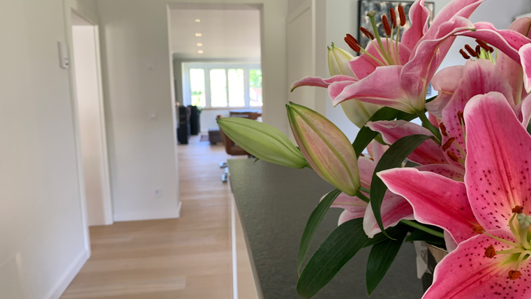 Een lichte binnenruimte met een houten vloer en grote ramen. Op de voorgrond staat een bloemenvaas met roze lelies.