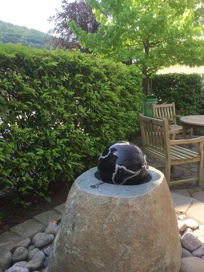 Eine ruhige Gartenlandschaft mit einem Stein und einer glänzenden Globus-Skulptur. Im Hintergrund sind ein Holzstuhl und ein grüner Busch zu sehen.