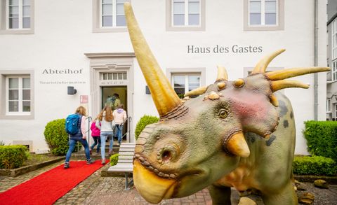 Een levendig dinosauriermodel staat voor de ingang van het "Huis van de Gast". Bezoekers lopen over een rode loper naar binnen.
