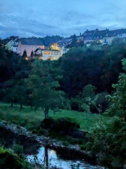 Un petit village au crépuscule, entouré d'arbres et de collines. Un petit ruisseau traverse le paysage, tandis que quelques maisons sont doucement éclairées.