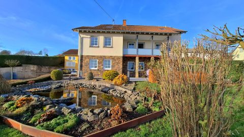 Ein zweigeschossiges Haus mit einem gepflegten Garten. Im Vordergrund ist ein kleiner Teich und bunte Pflanzen zu sehen.