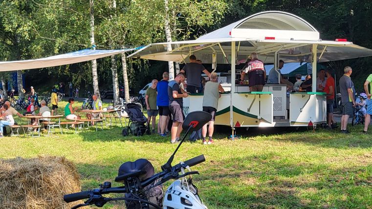 Ein belebter Imbissstand in der Natur, umgeben von Menschen, die essen und plaudern. Im Vordergrund steht ein Fahrrad mit Helm.
