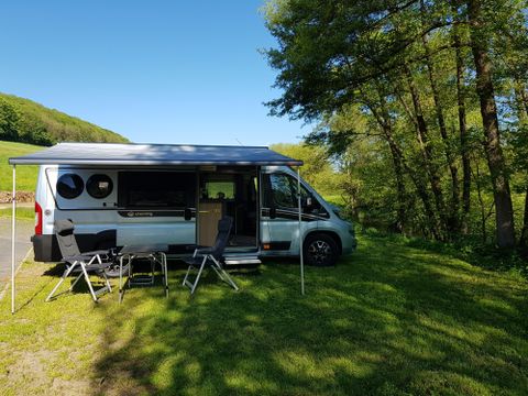 Un camping-car est garé sur une pelouse, entouré d'arbres. Deux chaises pliantes sont placées devant le véhicule, idéales pour se détendre dans la nature.