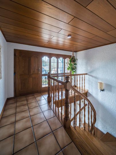 Ein einladender Eingangsbereich mit einer Holztreppe und einer hellen Wand. Die Tür aus Holz öffnet sich zu einem gut beleuchteten Raum mit Fliesenboden.