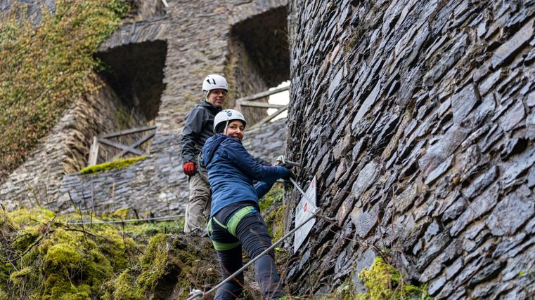 Zwei Personen beim Klettern an einer alten Steinmauer. Die Umgebung ist grün und bewaldet, mit einem historischen Gebäude im Hintergrund.