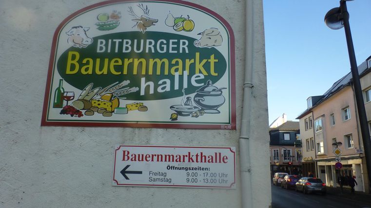 Ein Schild für die Bitburger Bauernmarkthalle mit bunten Illustrationen von Lebensmitteln. Darunter stehen die Öffnungszeiten des Marktes.