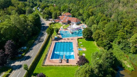 Ein Luftbild eines Schwimmbads mit mehreren Becken und einer Rutsche, umgeben von grüner Natur. Im Hintergrund ist ein Gebäude und eine Straße zu sehen.