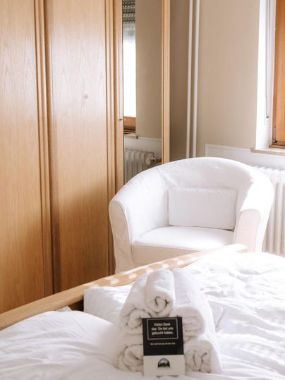 Ein gemütliches Schlafzimmer mit einem weißen Sessel und Holzschrank. Frisch gefaltete Handtücher liegen auf dem Bett.