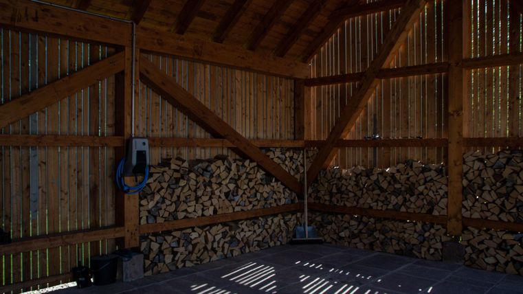 Ein Holzschuppen mit einer Holzstapelwand und einem offenen Dach. Sonnenstrahlen fallen auf den Boden und erzeugen ein interessantes Lichtspiel.