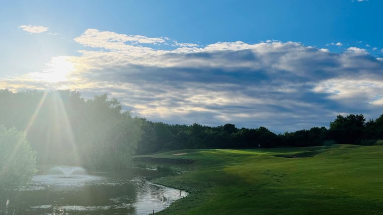 Eine grüne Golfplatzlandschaft mit sanften Hügeln und einem kleinen Teich. Der Himmel ist blau mit einigen Wolken und strahlendem Sonnenlicht.