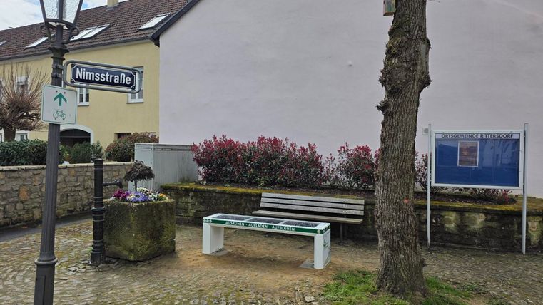 Eine ruhige Ecke mit einer Bank und einem Straßenname-Schild "Nimstraße". Im Hintergrund sind Blumen und ein Baum sichtbar, umgeben von gepflastertem Boden.
