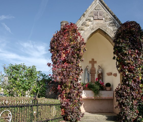 Small chapel with statues, surrounded by autumn ivy. A wrought-iron gate and blue sky in the background., © TI Bitburger Land