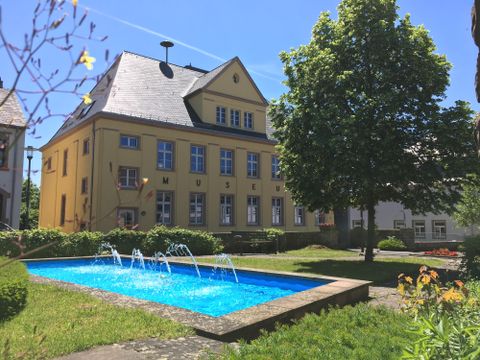 A yellow building stands in a green garden with a blue water basin. In the background, trees and a clear sky can be seen.