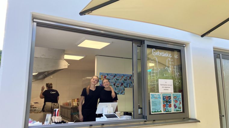 Two women are standing at a counter and smiling. In the background, kitchen appliances and colorful posters can be seen.