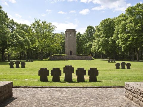 Eine weitläufige Grünfläche mit einem steinernen Turm in der Mitte. Rundherum stehen Steinblockskulpturen, umgeben von Bäumen und blauem Himmel.