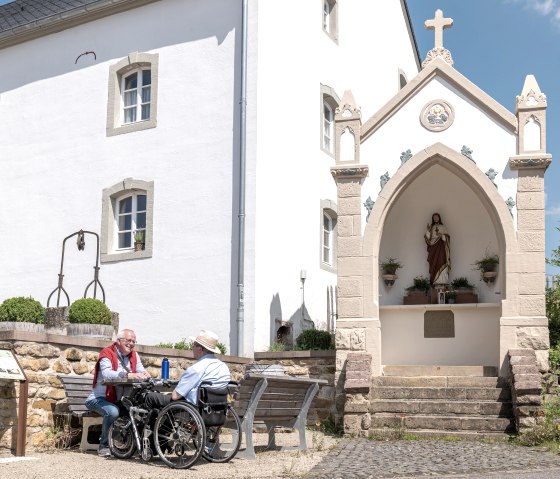Zwei M&auml;nner, einer im Rollstuhl, sitzen auf einer Bank vor einem wei&szlig;en Geb&auml;ude mit einer religi&ouml;sen Statue in einer Nische. Sonniger Tag., &copy; Naturpark S&uuml;deifel, Thomas Urbany