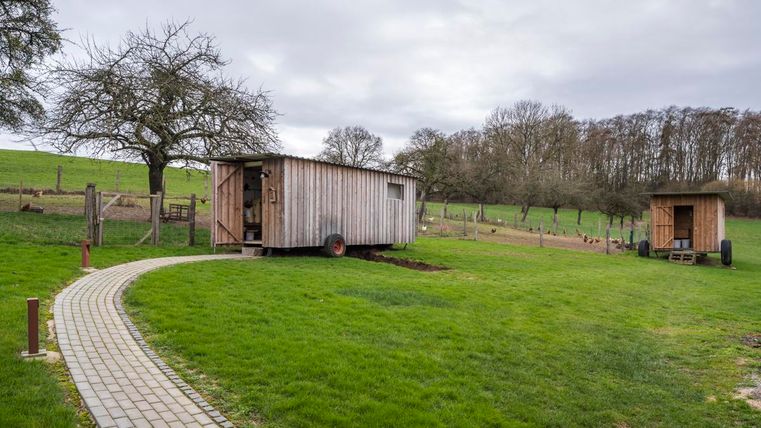 Eine idyllische Landschaft mit einem Holzhaus und einem kleinen Nebengebäude. Der Weg ist gepflastert und umgeben von grüner Wiese und Bäumen.
