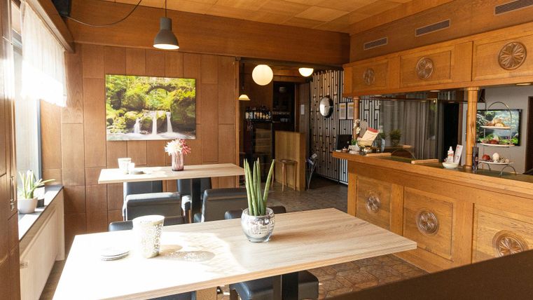 Ein gemütliches Restaurant mit Holzverkleidung und hellen Tischen. An der Wand hängt ein Bild von einem Wasserfall und es gibt Zimmerpflanzen auf den Tischen.