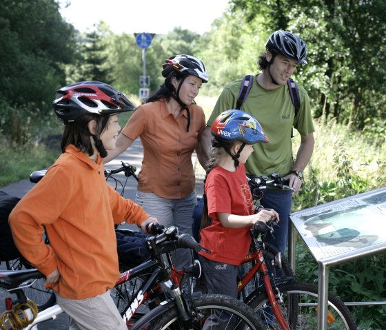 Familie mit Fahrr&auml;dern liest eine Infotafel &uuml;ber Biber auf dem Eifel-Ardennen-Radweg. Sie tragen Helme und sind von gr&uuml;ner Natur umgeben., &copy; Eifel Tourismus GmbH/intention