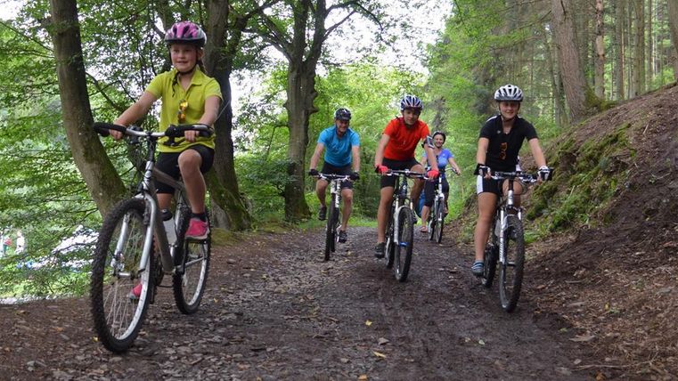 A group of cyclists is riding on a narrow, muddy forest path. The trees provide a green background and the riders are wearing helmets.