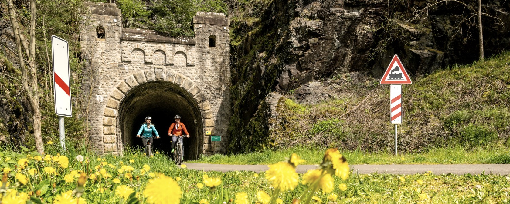 Zwei Radfahrer auf einem Weg vor einem alten Bahntunnel, umgeben von bl&uuml;hendem L&ouml;wenzahn und Verkehrsschildern., &copy; Eifel Tourismus GmbH, Dominik Ketz