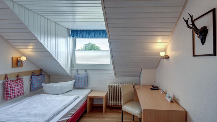 Une chambre confortable sous le toit avec un lit simple et un bureau. Une fenêtre laisse entrer la lumière du jour et offre une vue sur l'extérieur.