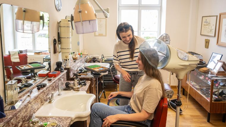 A hair salon with a customer sitting in a hairdresser's chair. The hairdresser is talking to her, and a bonnet is on the customer's head.