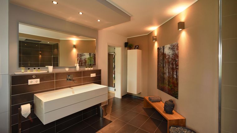 Une salle de bain moderne avec un lavabo blanc, un miroir et un éclairage doux. Les murs sont carrelés en sombre, et il y a un banc en bois ainsi qu'une photographie artistique au mur.