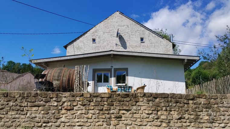 Een charmant huis met een lichte buitenkleur en een terras. Op de voorgrond staat een stenen muur en een houten vat.
