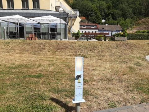 A bright, modern light fixture stands on a lawn in front of a building. In the background, tablecloths and umbrellas of an outdoor area are visible.