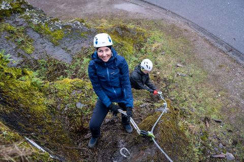 Eine Frau klettert mit einem Seil an einer steilen, moosbedeckten Wand. Im Hintergrund ist eine weitere Person, die ebenfalls klettert.