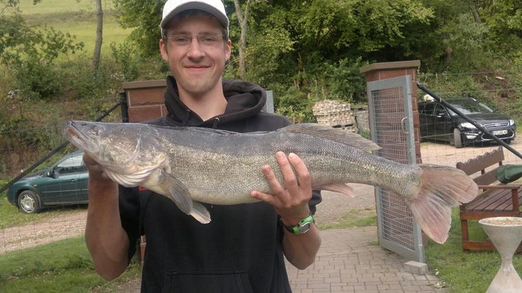 Un jeune homme tient un gros poisson dans ses mains. En arrière-plan, on voit des arbres et des voitures.
