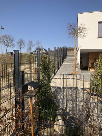 Ein moderner Gartenweg mit einem Metallzaun führt zu einem Haus. Im Hintergrund sind Bäume und ein blauer Himmel sichtbar.