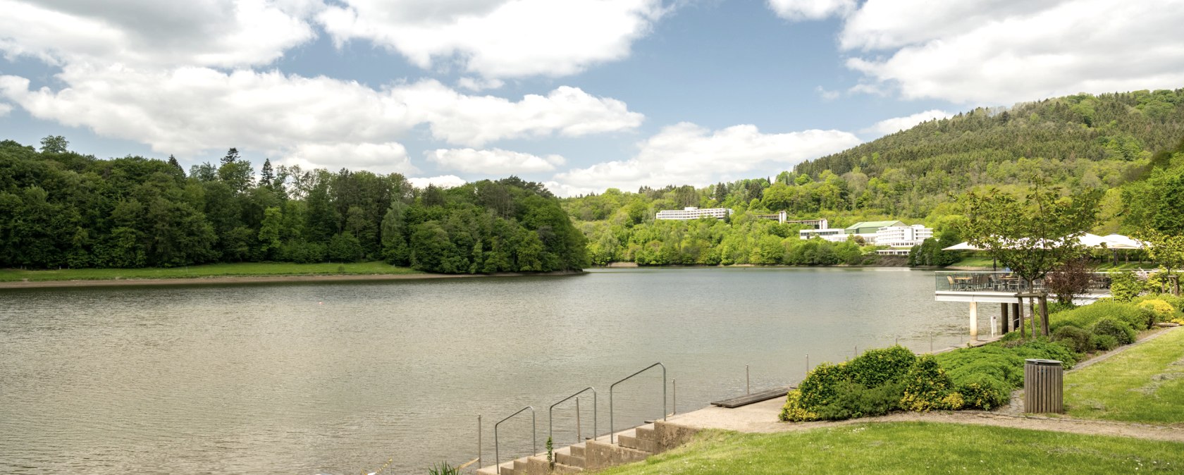 Stausee Bitburg mit gr&uuml;ner Uferlandschaft, B&auml;umen und Geb&auml;uden im Hintergrund unter einem bew&ouml;lkten Himmel., &copy; Eifel Tourismus GmbH, Dominik Ketz