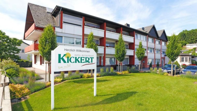 Un bâtiment moderne avec une enseigne accueillante "Bienvenue" se dresse au premier plan. Le jardin soigné est agrémenté d'arbres et d'herbe fraîche.