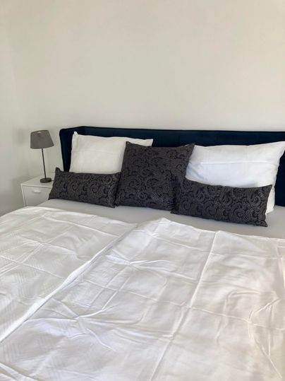 A modern bedroom with a large bed decorated with white sheets and several pillows. This is complemented by a nightstand with a small lamp.