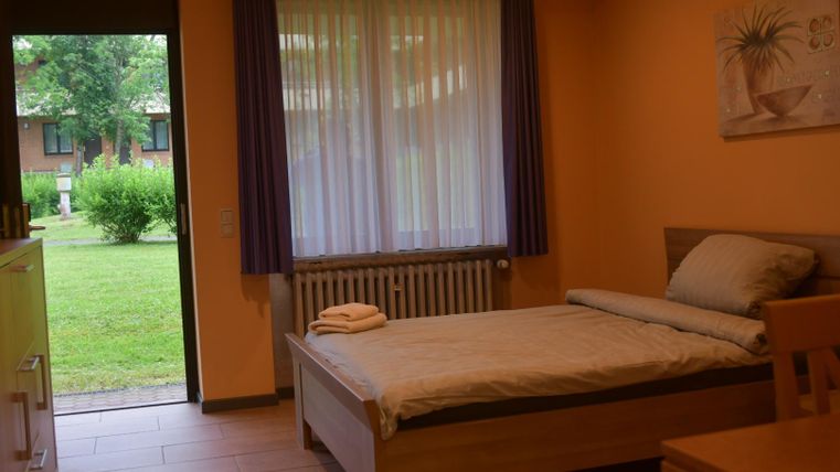 Een eenvoudige slaapkamer met een eenpersoonsbed en een tafel. Het uitzicht vanuit het raam toont een groene tuin.