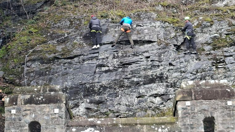 Drei Kletterer steigen eine Felswand hinauf. Im Vordergrund ist ein historische Mauer aus Stein zu sehen.