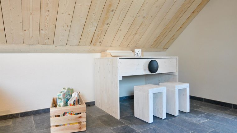 Ein modernes Zimmer mit einem schlichten Tisch und zwei Hockern. In einer Ecke steht eine Kiste mit dekorativen Elementen.