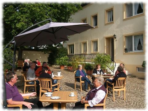 Eine Gruppe von Menschen sitzt gemütlich unter einem Sonnenschirm auf einer Terrasse. Es gibt Tische mit Kaffeetassen und eine ruhige, einladende Atmosphäre.