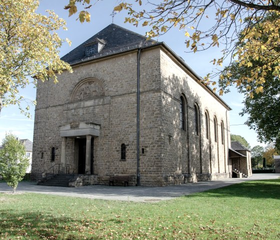 Die Kirche St. Hubertus in Wolsfeld, aus hellem Stein gebaut, steht in einer herbstlichen Umgebung mit Bäumen und grünem Rasen., © Tourist-Information Bitburger Land