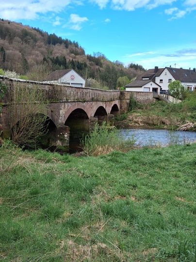 Un ancien pont en pierre sur une petite rivière, entouré de prés verts et d'arbres. En arrière-plan, on peut voir quelques maisons et un ciel bleu clair.
