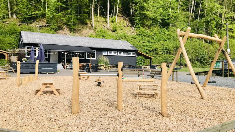 Ein Spielplatz mit Holzspielgeräten und Sitzgelegenheiten. Im Hintergrund befindet sich ein modernes Gebäude, umgeben von Bäumen.