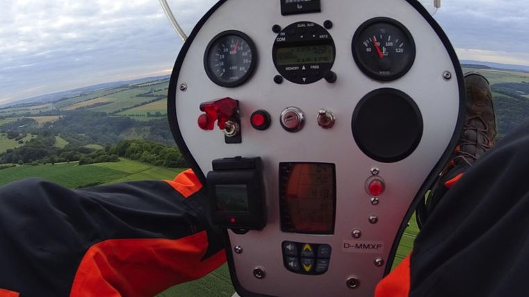 A view of the cockpit of a glider with various instruments. In the background, green fields and the sky are visible.