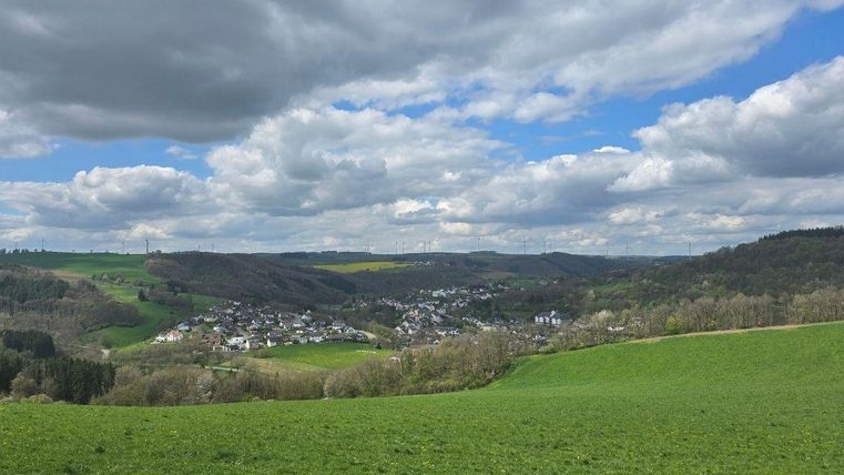 Een groene heuvelachtige landschap met een klein dorp in de dalen. De lucht is bewolkt en toont enkele zonnige plekken.