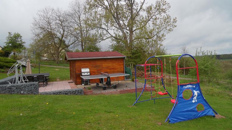 Een speeltuin met verschillende speeltoestellen, waaronder een schommel en een glijbaan. Op de achtergrond bevindt zich een houten huis en enkele bomen.